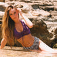 Woman in a purple top and checkered shorts sitting on rocks by a water source