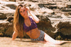 Woman in a purple top and checkered shorts sitting on rocks by a water source